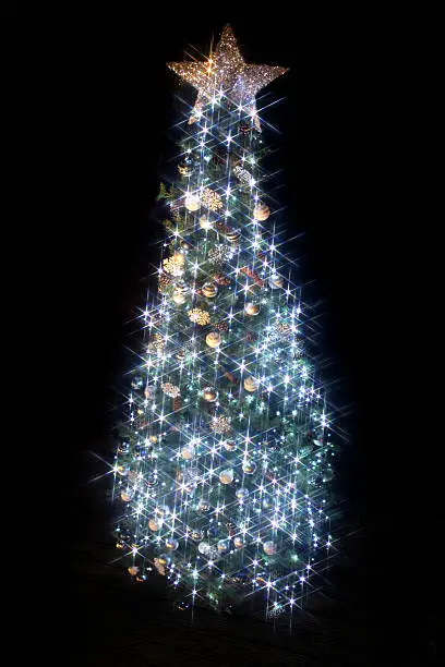 Photo showing an image of an artificial Christmas tree decorated with white LED fairy lights sparkling, isolated against a black night background. This image is taken through a star filter and shows six point stars. The tree is covered with themed Christmas decorations, including blue and white baubles, silver snowflakes, and pine cones, which have been wired onto the branches and needles of the fake tree. The tree is topped with a Christmas star.