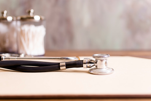 Close-up view of a black and silver doctor's stethoscope on a wooden desk in doctor's office.  Cotton swabs in jars in background.  No people.  Medical, healthcare, check-up, exam, cardiologist themes.