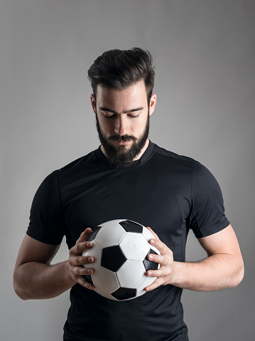 Intense portrait of football player holding and looking at the ball focused over gray studio background. Determination concept.