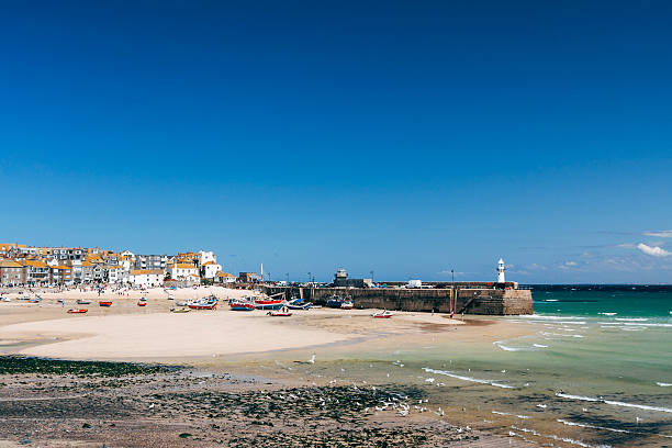 The picturesque St Ives harbour and beach at low tide in Cornwall, England. Small fishing boats are moored to the sand, historic buildings of St Ives can be seen lining the beach and quayside. The stone pier has a small lighthouse on it.