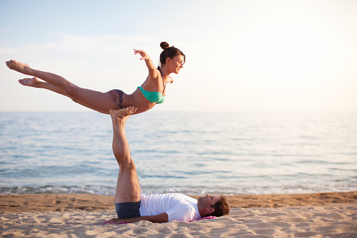 Young woman doing acro yoga with man on the beach by the sea. They are enjoying in exercise and stretching at sunset and beautiful light