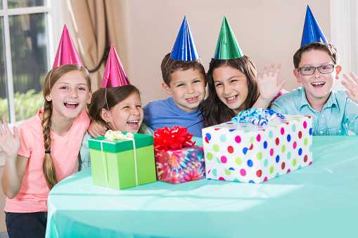 A group of five multi-ethnic children at a birthday party wearing party hats. Gifts in boxes, wrapped and topped with bows are on the table where they are sitting. They are waving, smiling and looking at the camera. The three children in the middle are Hispanic.