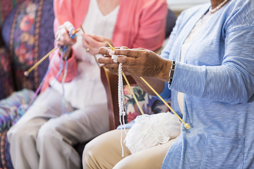 Cropped view of two unrecognizable multi-ethnic senior women sitting in the living room, knitting. The focus is on hands of the African American woman in the foreground.