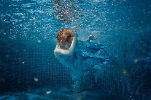 Women with long brown hair and white swimsuit diving underwater after jumping in the sea, surrounded by air bubbles, view from the side