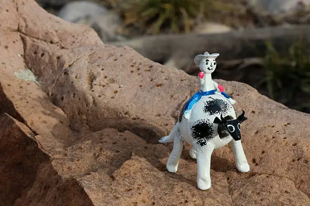 Photograph of a smiling toy coyboy riding a black and white bobble head cow.  The small wooden toy is sitting on a light brown rock.  The bobble head toy is hand painted and appears to be waving at the camera.  The smiling, waving toy gives a feeling of whimsy and playfulness.