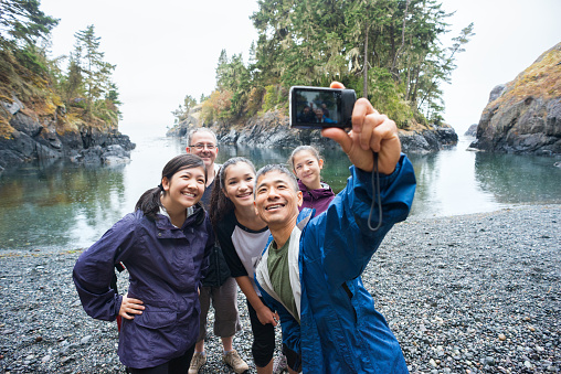A real extended family of multi-ethnic and multi-generational backpackers in the wilderness pose of a selfie on a deserted beach surrounded by islands and forest with the ocean in the background.  Rainy day in a wilderness park.  Whiffen Spit, Sooke, British Columbia, Canada.