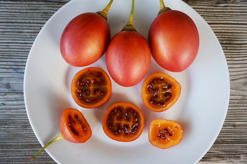 Whole and sliced fresh Tamarillo fruits on a white plate on wooden background.