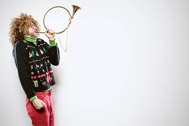 Christmas Sweater Man with Trumpet A man in a knit ugly Christmas cardigan button up sweater, complete with matching red pants and a green turtleneck, blows into a Christmas trumpet.  Horizontal with copy space. man trumpet stock pictures, royalty-free photos & images