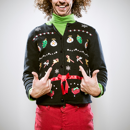 A man in a knit ugly Christmas cardigan button up sweater, complete with matching red pants and a green turtleneck.  He points proudly to his overly decorated sweater, a smile on his face.  Square crop.