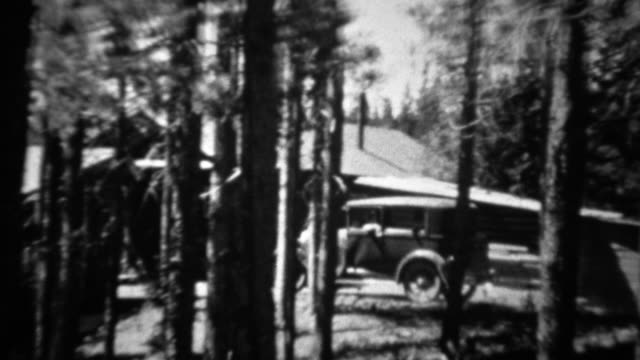 1934: Ford model A car driving past log cabin thru pine tree forest.