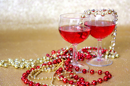 Two wine glasses filled with a red drink are surrounded by bright beaded New Years Eve necklaces in front of a glittery gold background