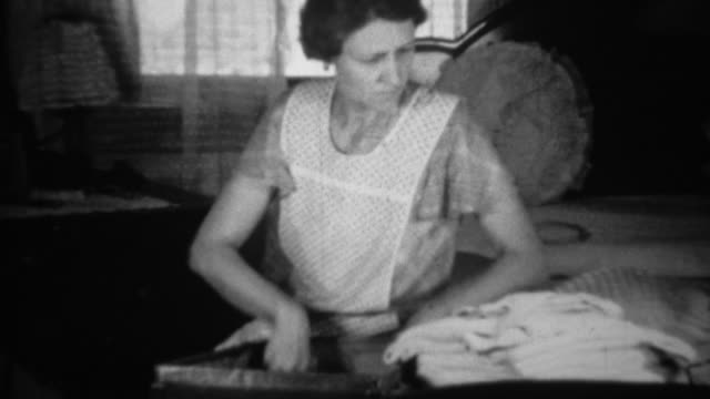 1936: Woman packing clothes into suitcase seated in living room.