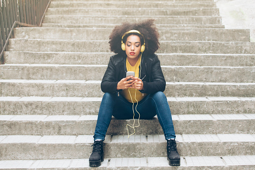 Young attractive girl in urban background listening music with yellow headphones sitting on stairs. Wear yellow pullover, black jacket and blue jeans. Holding phone.