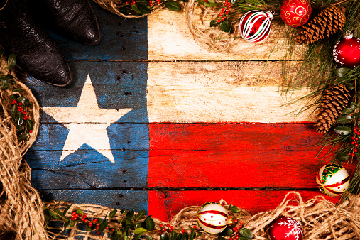Happy Southern Holidays!  Rustic Texas State flag craft created from old wooden boards and painted red, white, and blue.  Christmas decorations: ornaments, pine cones, pine branches, holly, cowboy boots, garland, burlap surround flag to form a frame.  Copyspace in center.