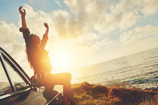 Cropped shot of a young woman sitting on the hood of her car while on a road trip