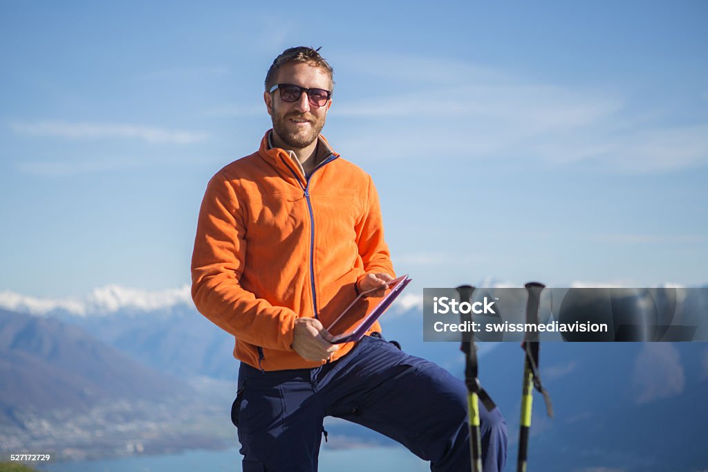 Technology at top of mountain Young man hiking and wearing sport clothing standing on bench using a digital tablet.Mountain lake landscape of Switzerland. Ticino Canton. Digital Tablet Stock Photo