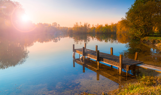 A wooden jetty juts out onto a calm lake with autumn coloured trees bordering the shores. The sun is setting behind the trees on the far bank