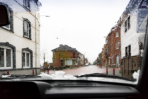 Look through the windshield of a car with some snow and rain. The car is driving through a quiet street in a small town in Belgium