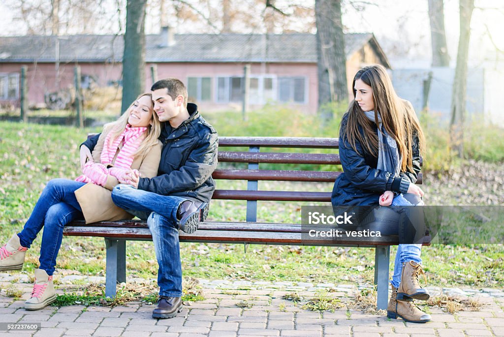 Really hate you A couple of young man and woman teenagers sitting on a bench, embracing, in love and happy at the other end sits a sad alone girl Couple - Relationship Stock Photo