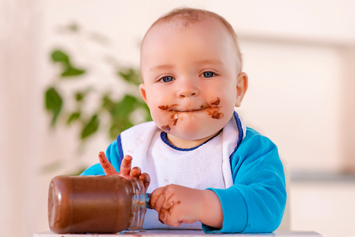 A baby grabbing a jar of chocolate cream while sitting in his high chair.His face and hands smeared with chocolate cream.