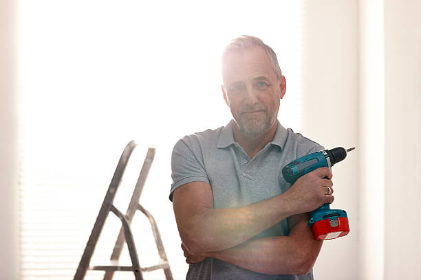 Mature man at home with a drill machine Portrait of mature man standing in living room holding a drill machine holding drill stock pictures, royalty-free photos & images