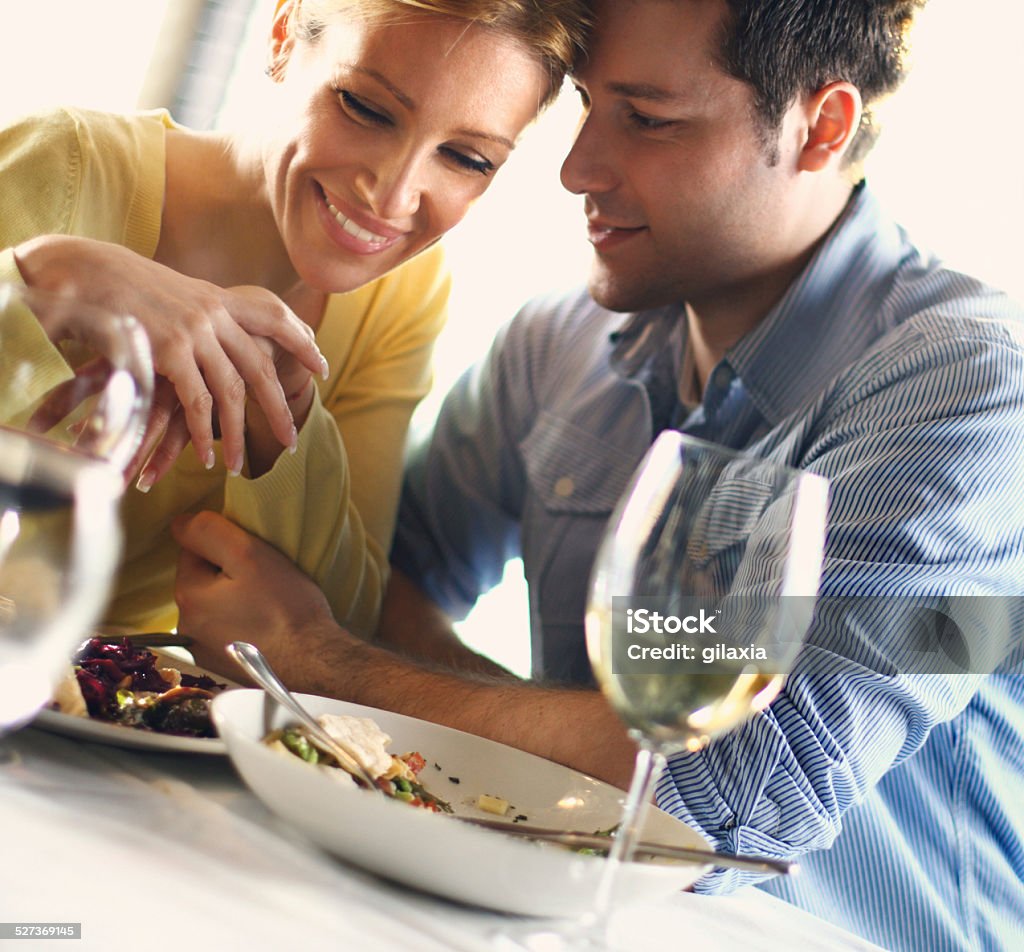 Couple having romantic dinner. Young to middle aged couple having romantic meal in a restaurant.Their head are touching.She's smiling while he's whispering something to her ear.Wearing yellow long sleeved shirt.Her hair is dark blond,pulled back with a clip.He's also smiling,wearing casual blue shirt.They're having glass of wine with food.Tilt,indoors. 20-29 Years Stock Photo