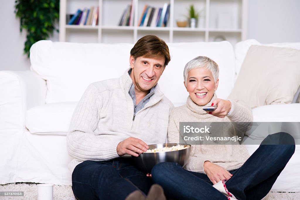 Mature couple watching TV Mature couple watching TV, eating pop corn and sitting on carpet at living room. 40-44 Years Stock Photo