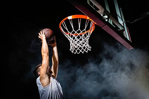 Young basketball player in white jersey dunking the ball in the basket. Foggy foreground, black background.