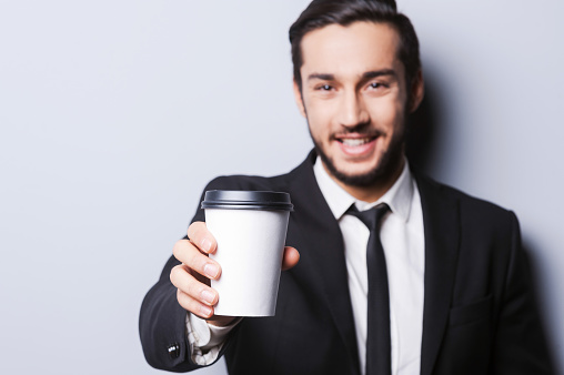 Portrait of confident young man in formalwear looking at camera and stretching out a cup of coffee while standing against grey background
