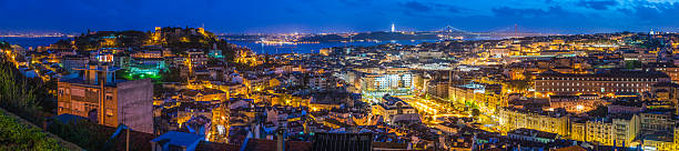 Lisbon central cityscape panorama landmarks waterfront illuminated at dusk Portugal The landmarks of Lisbon illuminated by the warm glow of street lights under deep blue dusk skies, from the historic battlements of Castelo de San Jorge over the plazas of Baixa, the Elevador de Santa Justa and villas of Bairro Alto to the lights of the 25 de Abril Bridge and the outstretched arms of Cristo-Rei across the River Tagus, Portugal. ProPhoto RGB profile for maximum color fidelity and gamut. santa justa lift portugal stock pictures, royalty-free photos & images