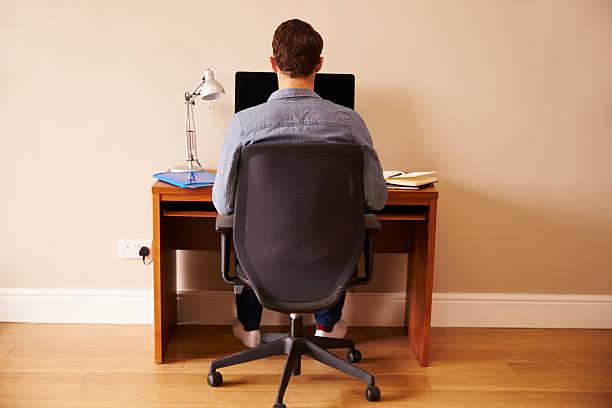 Man Sitting At Desk Working At Computer In Home Office Rear View Of Man Sitting At Desk Working At Computer In Home Office man in the desk back view stock pictures, royalty-free photos & images