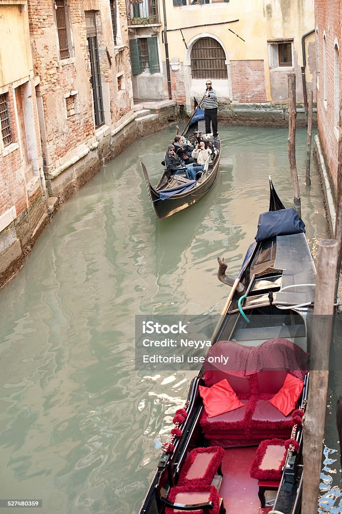 Tourism in Venice Venice, Italy - February 21, 2012; Canal in Venice with empty red decorated gondola at the wall and another with gondolier and tourists who enyoing beautiful day in Venice, Italy. Canal Stock Photo