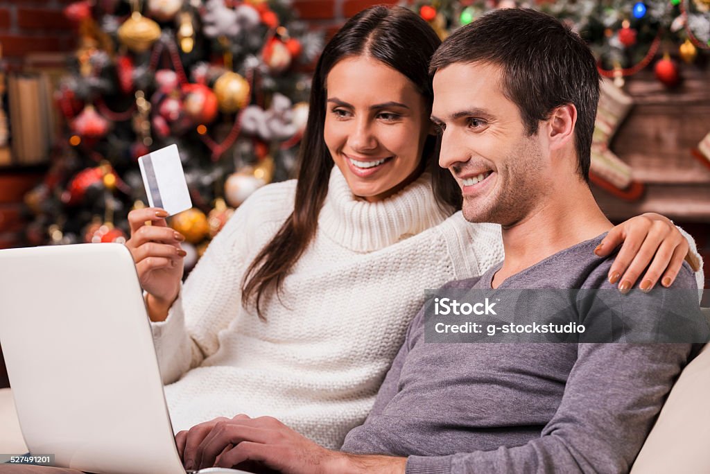 Taking advantages of Christmas sales. Beautiful young couple buying online while using computer together with Christmas Tree in the background Christmas Stock Photo