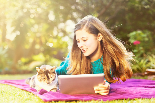 A pretty young woman lies on a blanket in a garden, holding her digital tablet, her cat next to her. The cat is taking her attention away from the tablet, but she doesn't mind! It's the perfect way to spend an afternoon! Copy space  on the shrubbery behind her.