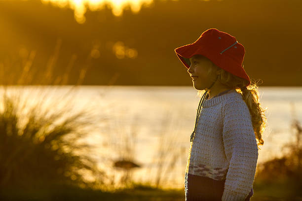 close up of a four year old child wearing a bright red hat  standing on the shore of a lake at sunset