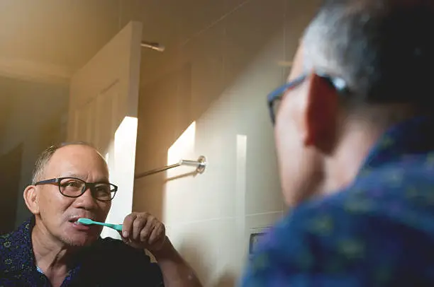 Asian oldman is Brushing Teeth and looking through the mirror in the bathroom.
