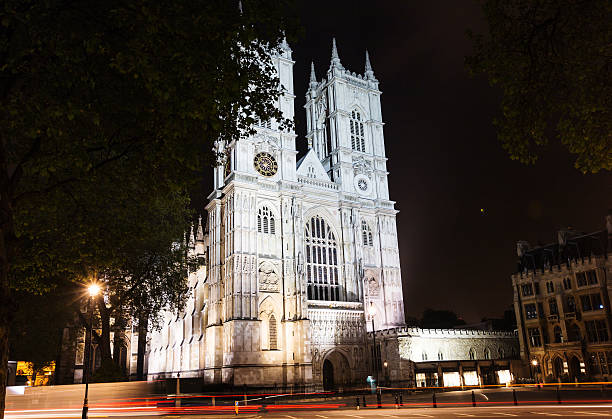 Bath Abbey is illuminated at night in Bath, Somerset, England, UK. The Gothic church facade features a large stained-glass window, sculptural decoration, and vertical stonework characteristic of late medieval ecclesiastical architecture. The abbey stands within the historic center of the city.