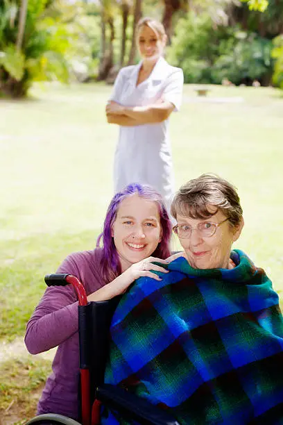 A young woman, possibly a granddaughter, embraces an old woman in a wheelchair who gives a mischievous smile as her nurse looks on, also smiling, from the background, as they all spend some time in a park.