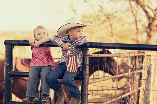 Two young children and siblings pose with the American flag on their farm in Texas. Cowboys and cowgirls.