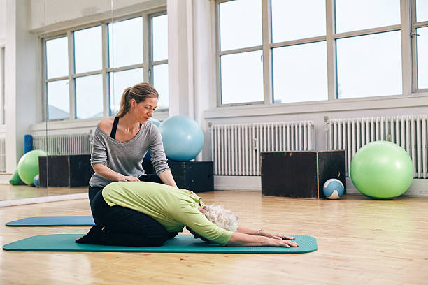 Female trainer helping senior woman doing yoga Female trainer helping senior woman doing yoga. Elder woman bending over a exercise mat with personal instructor helping at gym. childs pose stock pictures, royalty-free photos & images