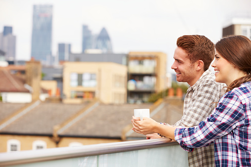 Couple Relaxing Outdoors On Rooftop Garden Drinking Coffee. Smiling Off Camera