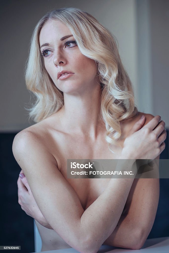 Slim Blonde Young blonde woman posing topless indoors. She has slim figure and long wavy hair. Sitting at the table. Adult Stock Photo