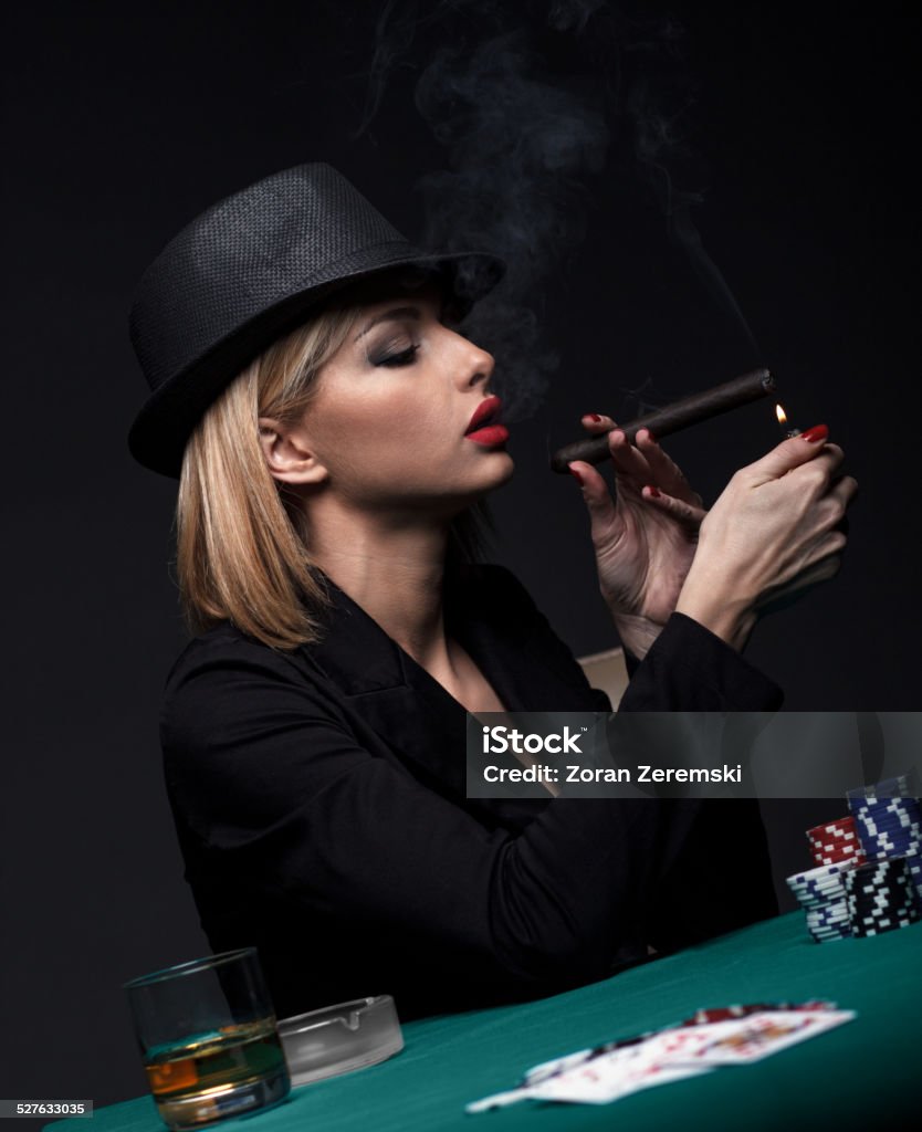Beautiful young woman playing poker Beautiful young woman lights a cigar during a poker game.Dark color Intensity. Whiskey Stock Photo
