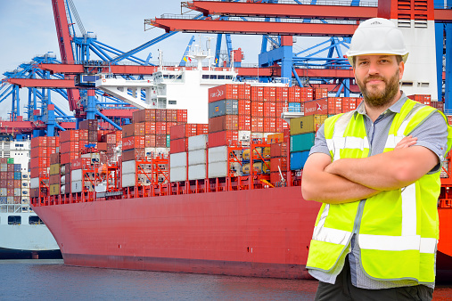 Dockworker standing in front of a container terminal in port. The man is wearing protective workwear and looking into the camera.