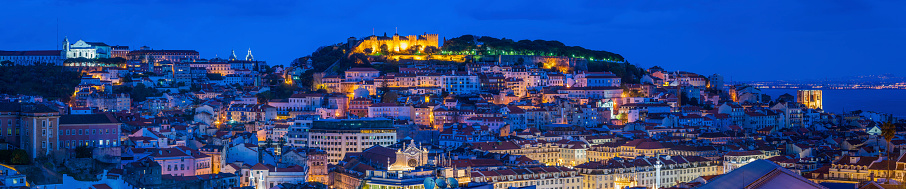 The iconic battlements of Castelo de Sao Jorge overlooking the rooftops of Lisbon illuminated by the warm glow of street lights under deep blue dusk skies. ProPhoto RGB profile for maximum color fidelity and gamut.