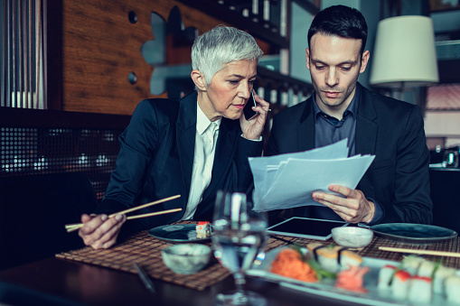 Businesspeople sitting in restaurant and having lunch. They are eating asian food. Mature woman talking on the phone and her colleague reading, finishing paperwork. Talking to client and have meeting.