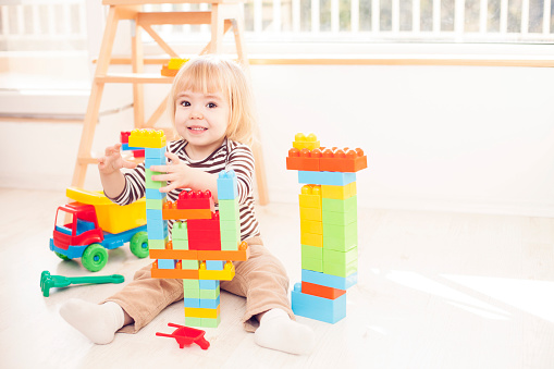 Little blond boy playing with lots of colorful plastic blocks in a sunny nursery room. Having fun, building and creating.