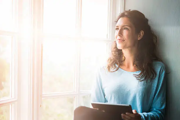 A photo of thoughtful woman sitting on window sill. Young female is holding digital tablet while looking away. She is wearing casuals in brightly lit room.