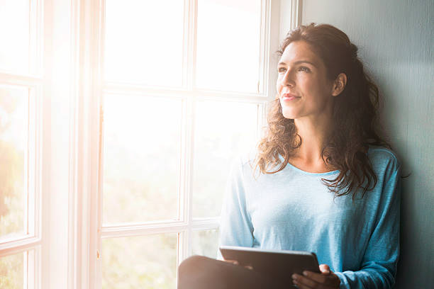 zamyślony młoda kobieta trzymająca cyfrowy tabletka przez okno - women reflection thinking window zdjęcia i obrazy z banku zdjęć