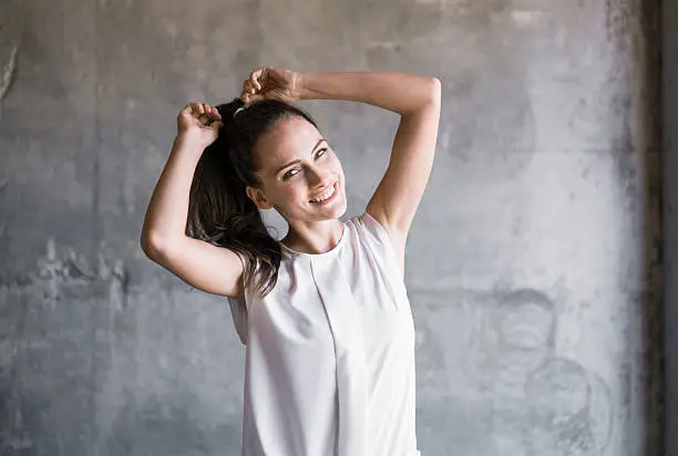 A photo of happy woman tying ponytail. Portrait of smiling young female is in casuals. She is standing against wall.